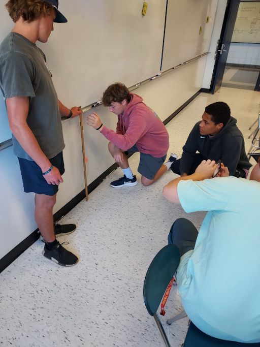 Four students in a classroom. One is holding a meter stick against the wall while another drops a bouncy ball in front of the meter stick. One student is watching while another takes a slow motion video of the ball.